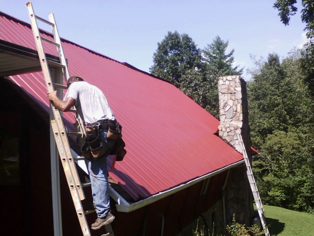 Roofer on a ladder beside a red metal roof and stone chimney — Soffit & Fascia Repair by Roofing Brothers in Silver Spring